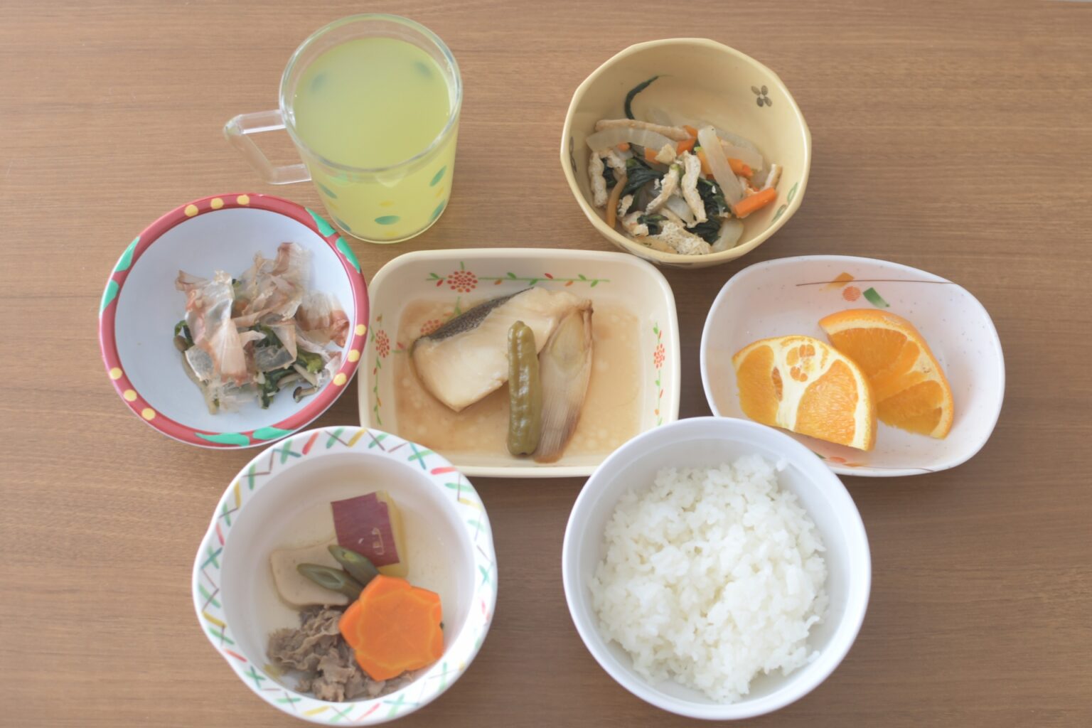 介護施設の食事風景。複数の小鉢に分けられた栄養バランスの良い食事が木製テーブルに配置されている。