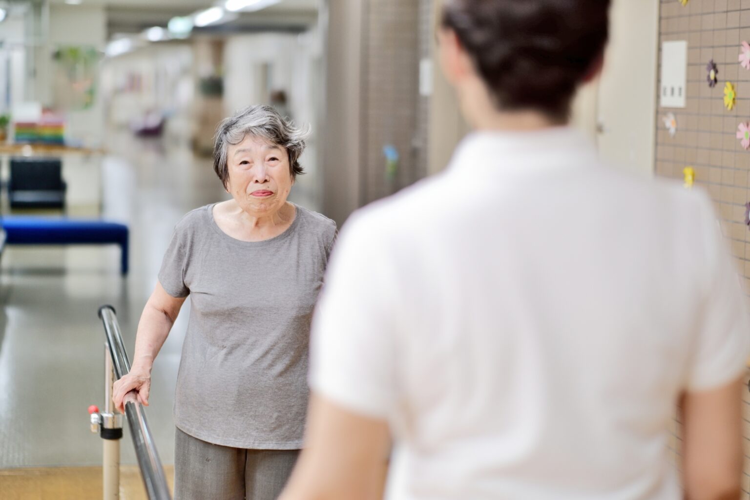 介護職員と高齢入居者が廊下で会話・交流している様子。リハビリ支援の場面。